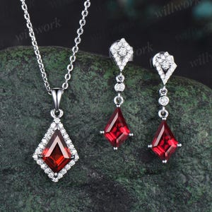 May include: A silver necklace and earrings set with a red gemstone pendant and matching earrings. The pendant and earrings are set in a diamond-shaped frame with smaller diamonds surrounding the gemstone.