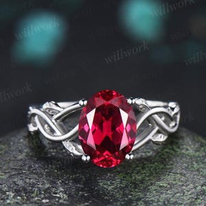 May include: A silver ring featuring a large, oval-cut, deep red gemstone set in the center. The band of the ring is crafted with an intricate, interwoven metal design that wraps around the finger, providing a detailed and textured appearance.
