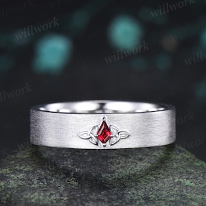 May include: A silver-toned band with a brushed matte finish featuring a central kite-shaped red gemstone. The stone is flanked by intricate metal knotwork detailing on both sides, creating a symmetrical design across the front of the ring.