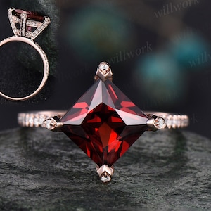 May include: A rose gold engagement ring with a square-shaped red gemstone surrounded by smaller diamonds. The ring has a pave setting with diamonds on the band.