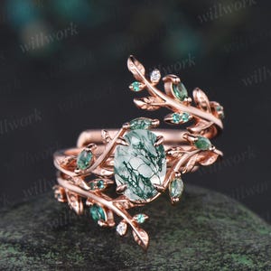 May include: Rose gold ring featuring an oval-cut, moss agate gemstone. The ring is adorned with leaf and vine detailing, accented with small green gemstones and tiny white stones. The design is nature-inspired and elegant.