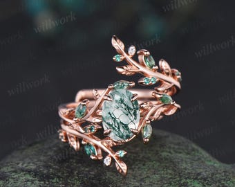 May include: Rose gold ring featuring an oval-cut, moss agate gemstone. The ring is adorned with leaf and vine detailing, accented with small green gemstones and tiny white stones. The design is nature-inspired and elegant.