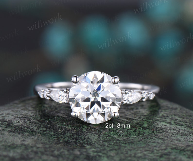 May include: A silver engagement ring featuring a large round centre stone and two pear-shaped accent stones. The centre stone is 2 carats and 8 millimetres in diameter. The ring is set on a silver band with small round stones.