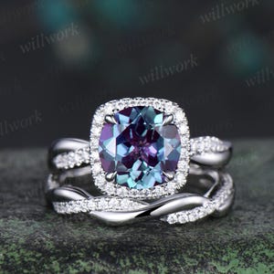 May include: A two-piece engagement ring set. The ring features a large, square-cut, color-changing gemstone surrounded by a halo of small diamonds. The band is a twisted design, also adorned with small diamonds. The ring is set in a silver metal.