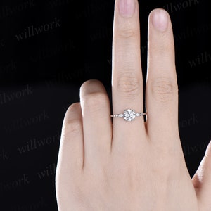 May include: A silver engagement ring with a round centre stone and smaller accent stones on the band. The ring is on a person's finger.