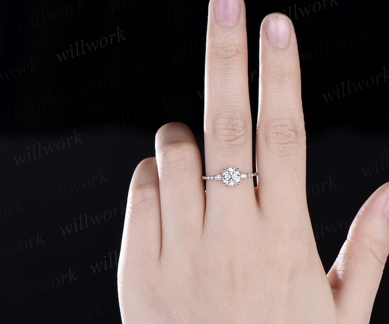 May include: A silver engagement ring with a round centre stone and smaller accent stones on the band. The ring is on a person's finger.