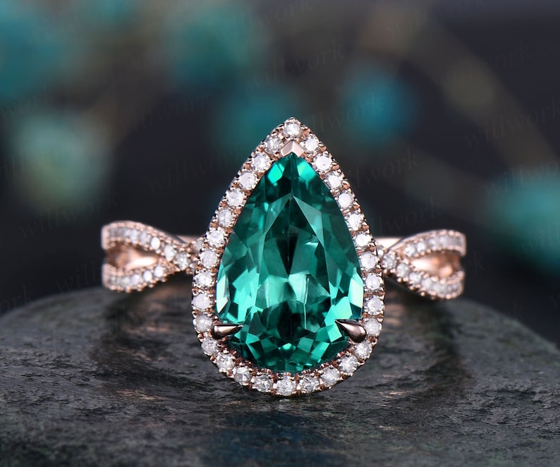 May include: A pear-shaped emerald green gemstone engagement ring with a halo of smaller diamonds set in rose gold. The ring features a delicate, twisted band with pave diamonds.
