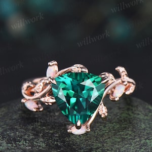 May include: Rose gold ring featuring a large, heart-shaped, emerald-green gemstone. The ring has delicate leaf and opal accents. The ring is set on a dark, textured surface.
