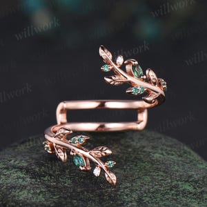May include: Rose gold ring featuring a leaf design. The leaves are embellished with small, emerald-green gemstones. The ring has an open design, with the leaves wrapping around the finger. The ring is displayed on a dark, textured surface.