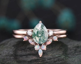 May include: A rose gold engagement ring set with a pear-shaped green moss agate gemstone surrounded by a halo of smaller white gemstones and a delicate band of opal gemstones.
