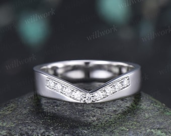 May include: A silver-toned V-shaped band ring featuring a row of small, clear, round-cut stones embedded along the center chevron design. The metal has a smooth, polished finish, and the ring is positioned on a textured, dark gray surface.