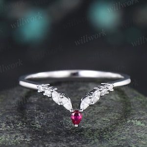 May include: A V-shaped ring crafted from polished silver-toned metal, featuring a central pear-cut red gemstone flanked by marquise-cut and round-cut clear stones arranged in a symmetrical chevron pattern along the band's upper curve.