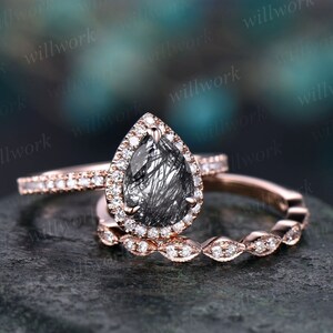May include: A trio of rose gold rings. The engagement ring has a pear-cut black gemstone, surrounded by small diamonds. The wedding band is a thin band of small diamonds, and the third ring has a repeating diamond pattern.