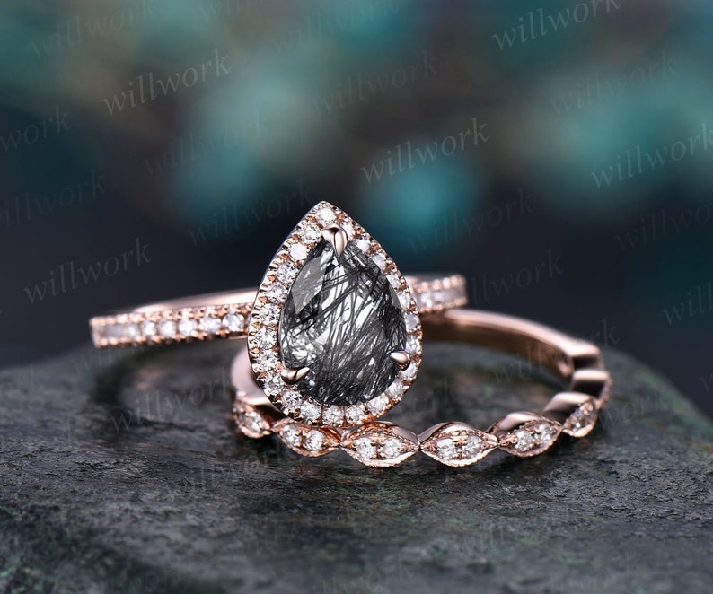 May include: A trio of rose gold rings. The engagement ring has a pear-cut black gemstone, surrounded by small diamonds. The wedding band is a thin band of small diamonds, and the third ring has a repeating diamond pattern.