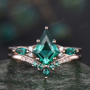 May include: A rose gold engagement ring set with a large emerald green square-shaped gemstone surrounded by smaller diamonds and emerald green stones. The ring is set on a delicate band.
