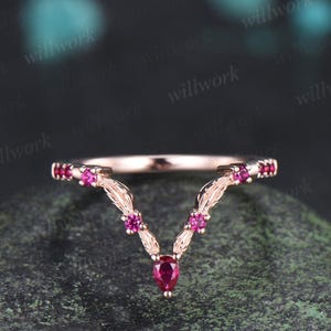 May include: A rose gold ring with a V-shaped design. The ring features small, round, and pear-shaped red gemstones. The band is adorned with delicate leaf-like details, creating an elegant and sophisticated look. The ring is set against a dark background.