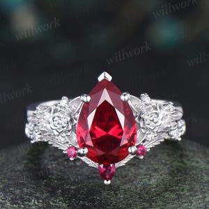 May include: A silver ring with a large, pear-shaped, deep red gemstone as the focal point. The ring features intricate floral detailing on the band and smaller red accent stones. The design is elegant and eye-catching.