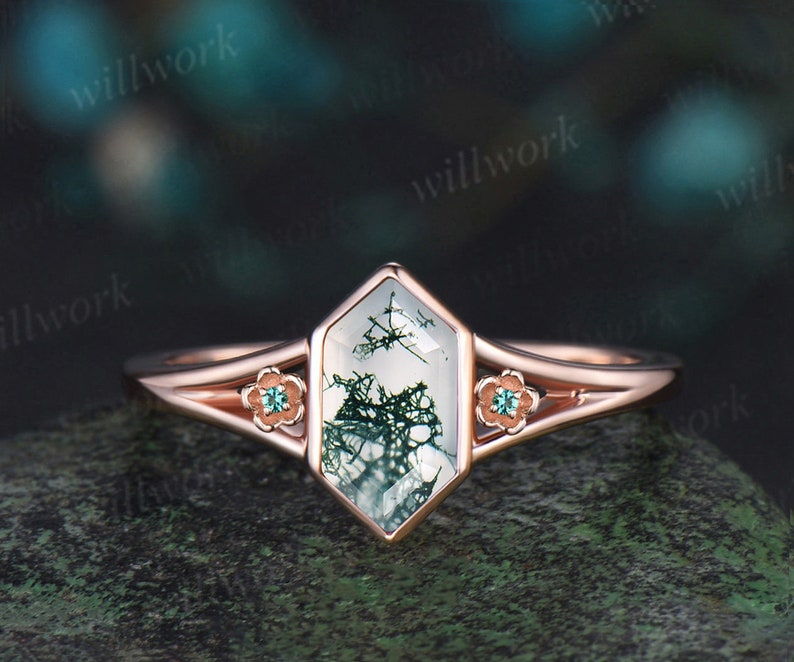 May include: A rose gold engagement ring with a hexagonal moss agate gemstone. The gemstone has a green and white pattern. The ring has two small green gemstones on each side of the main stone.