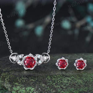 May include: A silver necklace and matching stud earrings set featuring round, faceted red stones set in floral-shaped metal mounts. The necklace pendant includes small white opal-like accents and clear stones along the silver metal leaves.