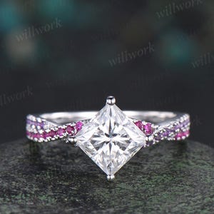 May include: A silver ring with a large, square, clear gemstone as the centerpiece. The band features alternating pink and purple gemstones. The ring is set against a dark, textured background.