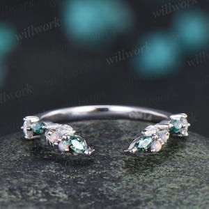 May include: A silver ring with a delicate design. The ring features small, leaf-like clusters adorned with teal and white gemstones. The gemstones are oval and round, adding a touch of sparkle. The ring is set against a dark, textured background.