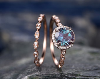 May include: A rose gold engagement ring set with a round alexandrite center stone surrounded by a halo of smaller diamonds. The matching wedding band features a delicate design of oval-shaped diamonds.
