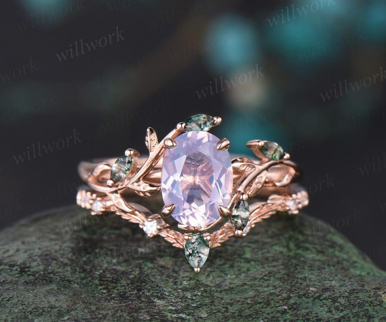 May include: Rose gold engagement ring and wedding band set. The engagement ring features a light pink oval-shaped gemstone, accented by smaller green gemstones and leaf-like details. The wedding band has small diamonds.  The set is designed with a nature-inspired theme.