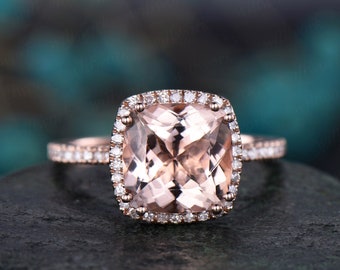 May include: A rose gold engagement ring with a cushion-cut morganite center stone and a halo of smaller diamonds. The ring has a delicate band with pave diamonds.