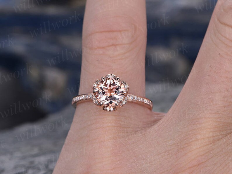 May include: A rose gold engagement ring with a round, light pink center stone surrounded by a halo of smaller diamonds. The band is also set with smaller diamonds.