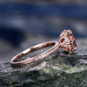 May include: A rose gold engagement ring with a round, light pink gemstone surrounded by smaller diamonds. The band is also set with small diamonds.