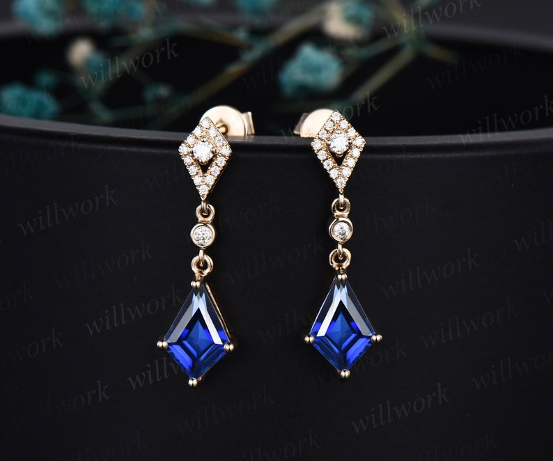 May include: A pair of gold dangle earrings with a square blue gemstone and a diamond halo setting. The earrings are on a black background.