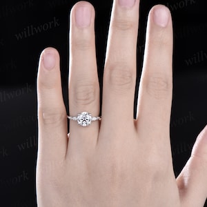 May include: A silver engagement ring with a round centre stone and smaller accent stones on the band. The ring is on a person's finger.