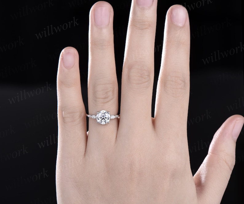 May include: A silver engagement ring with a round centre stone and smaller accent stones on the band. The ring is on a person's finger.