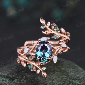 Peut inclure: Bague en or rose ornée d'une pierre précieuse ovale de couleur teal au centre. La bague présente des motifs de feuilles, de petites pierres blanches et vertes, et des détails fins. Le design est complexe et élégant.