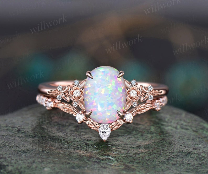 May include: A rose gold engagement ring with a large oval opal gemstone and smaller diamond accents. The ring is set with a delicate floral design and has a matching wedding band.