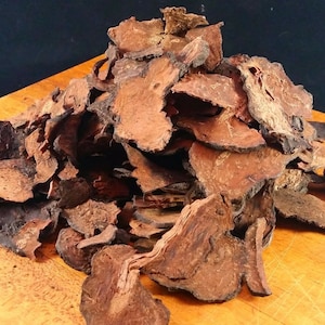 May include: A pile of dried, dark brown, irregularly shaped pieces, possibly bark or root, with a rough texture. The pieces are scattered on a light brown wooden surface, with a dark background.