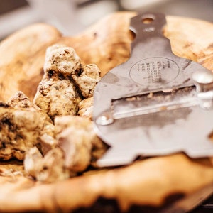 Op de afbeelding: Een houten kom gevuld met witte truffels en een zilveren truffelsnijder. De snijder heeft de woorden "TRUFFLE CLUB" gegraveerd.