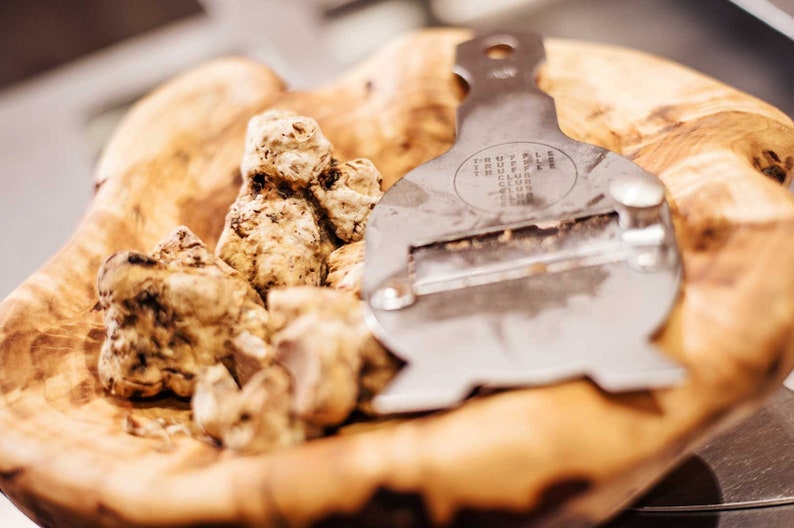 Op de afbeelding: Een houten kom gevuld met witte truffels en een zilveren truffelsnijder. De snijder heeft de woorden "TRUFFLE CLUB" gegraveerd.