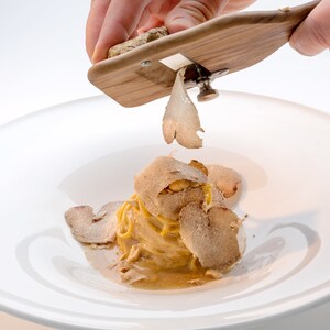 Op de afbeelding: Een hand raspt een truffel over een bord pasta met truffelraspels. De pasta ligt op een wit bord.