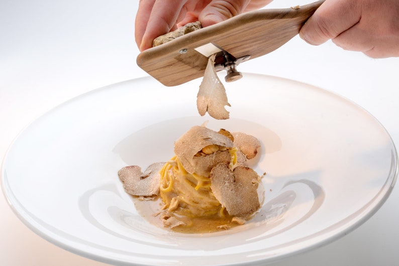 Op de afbeelding: Een hand raspt een truffel over een bord pasta met truffelraspels. De pasta ligt op een wit bord.