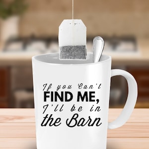 May include: White ceramic mug with black text that reads "If you can't find me, I'll be in the barn". A tea bag hangs from the rim of the mug.