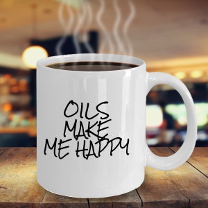 May include: White ceramic coffee mug with black text that reads "OILS MAKE ME HAPPY". The mug is filled with coffee and steam is rising from the mug.