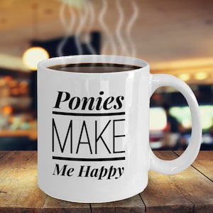 May include: A white ceramic mug filled with dark coffee and steam rising from the top. The mug features black text that reads Ponies MAKE Me Happy, with horizontal lines separating the words Ponies, MAKE, and Me Happy on the front.