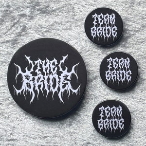 Könnte beinhalten: Drei schwarze und weiße Anstecknadeln mit dem Text "Team Bride" und eine schwarze und weiße Anstecknadel mit dem Text "The Bride" in einer gotischen Schrift.