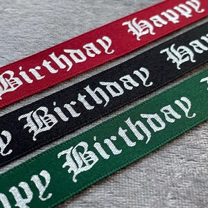 May include: Close-up of three ribbons in red, black, and green. Each ribbon has the words "Happy Birthday" printed in white, gothic-style lettering. The ribbons are likely for gift wrapping or crafting.
