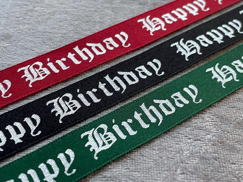 May include: Close-up of three ribbons in red, black, and green. Each ribbon has the words "Happy Birthday" printed in white, gothic-style lettering. The ribbons are likely for gift wrapping or crafting.