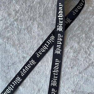 May include: Black ribbon with the words "Happy Birthday" in a gothic font. The ribbon is folded over itself, creating a loop. The background is a textured, light gray surface.