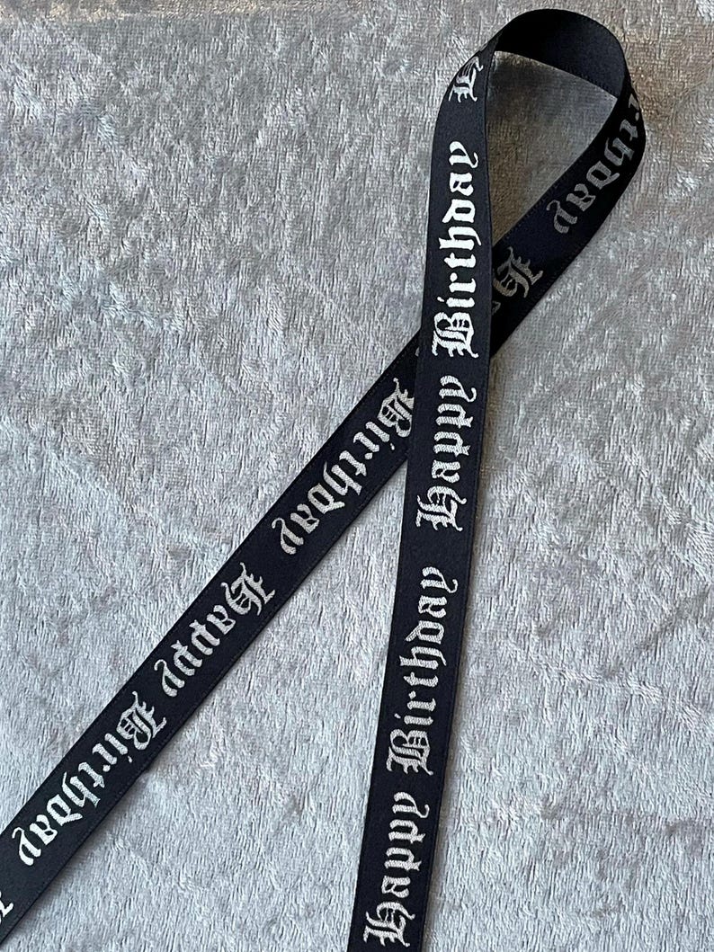 May include: Black ribbon with the words "Happy Birthday" in a gothic font. The ribbon is folded over itself, creating a loop. The background is a textured, light gray surface.