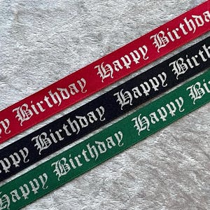 May include: Three ribbons in red, black, and green, each bearing the words "Happy Birthday" in a white, gothic font. The ribbons are set diagonally against a textured, light grey background.