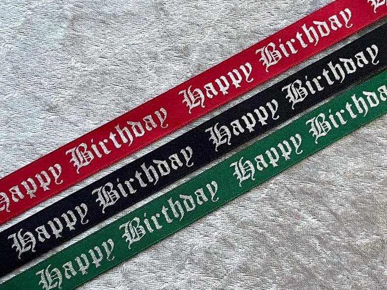 May include: Three ribbons in red, black, and green, each with the words "Happy Birthday" in a white, gothic-style font. The ribbons are diagonally arranged against a textured, light gray background.